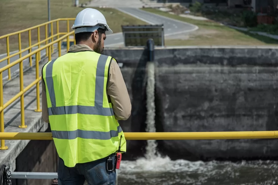 Worker in yellow vest and white hard hat looking down at water basin at wastewater treatment plant